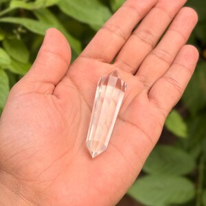 May include: A clear quartz crystal wand with a faceted, pointed tip. The crystal is held in an open hand against a backdrop of green foliage. The wand is approximately 4 inches long.