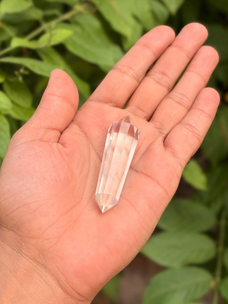 May include: A clear quartz crystal wand with a faceted, pointed tip. The crystal is held in an open hand against a backdrop of green foliage. The wand is approximately 4 inches long.