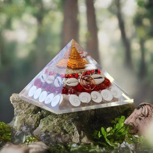 May include: A clear crystal pyramid with a gold pyramid inside. The pyramid is filled with red beads, white seashells, and a copper wire spiral.