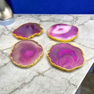 May include: Four purple agate coasters with gold trim. The coasters are round and have a natural, organic shape. They are perfect for adding a touch of elegance to any home.
