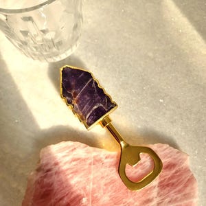 May include: A gold bottle opener with a purple amethyst stone on the top. The opener is sitting on a pink rose quartz.