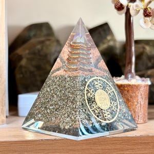 May include: A clear pyramid-shaped object with a gold-colored coil inside and a gold-colored circular emblem on the front. The pyramid is filled with dark gray and brown stones. The emblem has the numbers "303", "520", and "741".