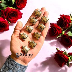 May include: A hand holds nine small, gold-coloured turtle figurines. The turtles are detailed with a textured shell and are arranged in a semi-circle. The background is a pink surface with red roses.