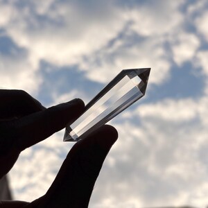 May include: A clear quartz crystal point held against a cloudy sky. The crystal is faceted and rectangular, with a pointed tip. The light shines through the crystal, creating a bright, transparent appearance. The crystal is held between two fingers.