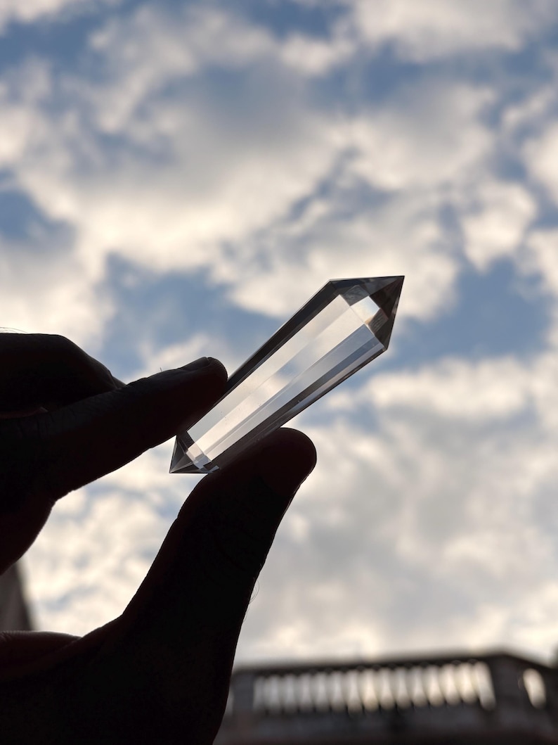 May include: A clear quartz crystal point held against a cloudy sky. The crystal is faceted and rectangular, with a pointed tip. The light shines through the crystal, creating a bright, transparent appearance. The crystal is held between two fingers.