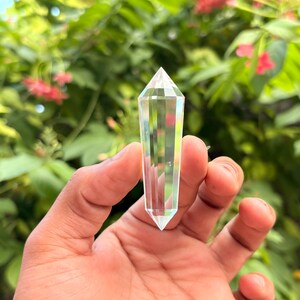 May include: A clear quartz crystal point held in a hand. The crystal is faceted and tapers to a point. The background is blurred green foliage with pink flowers. The crystal is a transparent, prism-like shape.