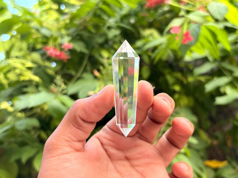 May include: A clear quartz crystal point held in a hand. The crystal is faceted and tapers to a point. The background is blurred green foliage with pink flowers. The crystal is a transparent, prism-like shape.
