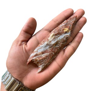 Puede incluir: Una punta de flecha de piedra roja y blanca. La punta de flecha mide aproximadamente 13 cm de largo y tiene una punta afilada.