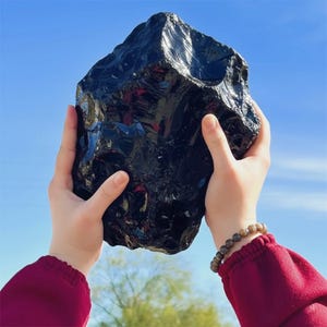 Puede incluir: Una gran piedra de obsidiana negra pulida sostenida contra un cielo azul. La obsidiana tiene una superficie brillante y reflectante y una forma irregular. La piedra está sostenida por dos manos, con una pulsera de cuentas visible en una muñeca.