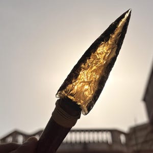 May include: A handcrafted arrowhead with a translucent, golden-brown blade. The blade is attached to a dark wooden handle wrapped with twine. The arrowhead is held up against the sunlight, highlighting its intricate texture and design.