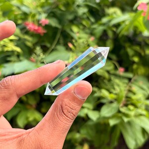 May include: A clear, faceted crystal wand held against a backdrop of green foliage and pink flowers. The crystal is rectangular with a pointed tip, showcasing internal striations and a subtle blue hue. The crystal is held between fingers.