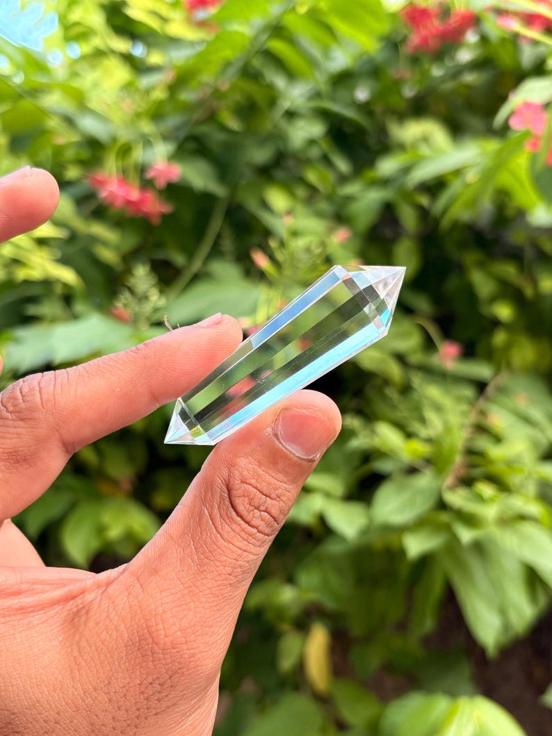 May include: A clear, faceted crystal wand held against a backdrop of green foliage and pink flowers. The crystal is rectangular with a pointed tip, showcasing internal striations and a subtle blue hue. The crystal is held between fingers.