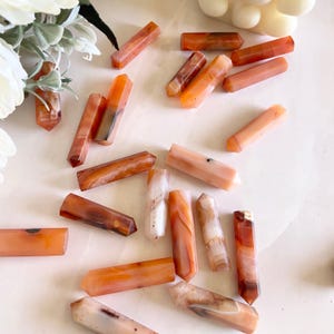 May include: A collection of carnelian crystal points in various shades of orange and white. The points are arranged on a white surface.
