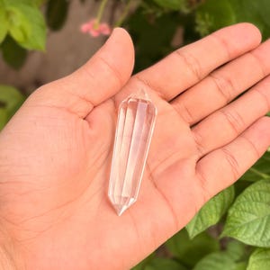 May include: A clear quartz crystal wand with a faceted design. The crystal is held in an open hand against a backdrop of green leaves and soft pink flowers. The wand is approximately 4 inches long.