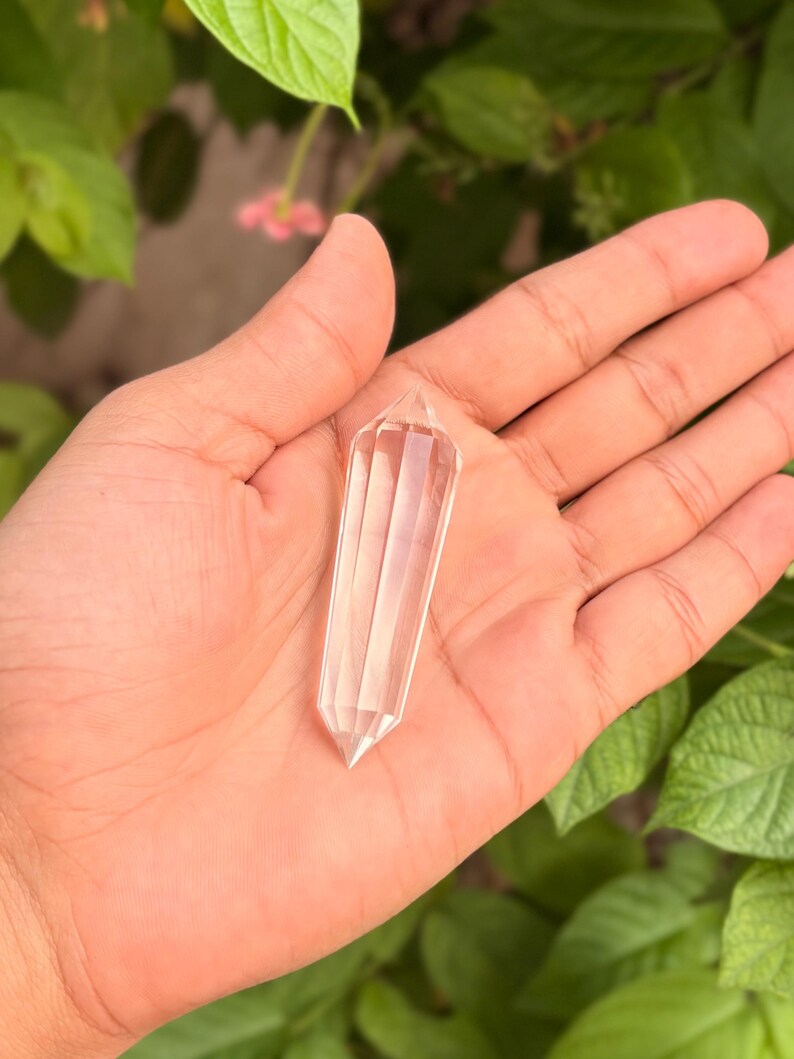 May include: A clear quartz crystal wand with a faceted design. The crystal is held in an open hand against a backdrop of green leaves and soft pink flowers. The wand is approximately 4 inches long.
