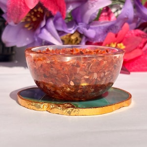 May include: A small, clear bowl filled with red, orange, and gold gemstone chips. The bowl is sitting on a green and gold agate coaster.