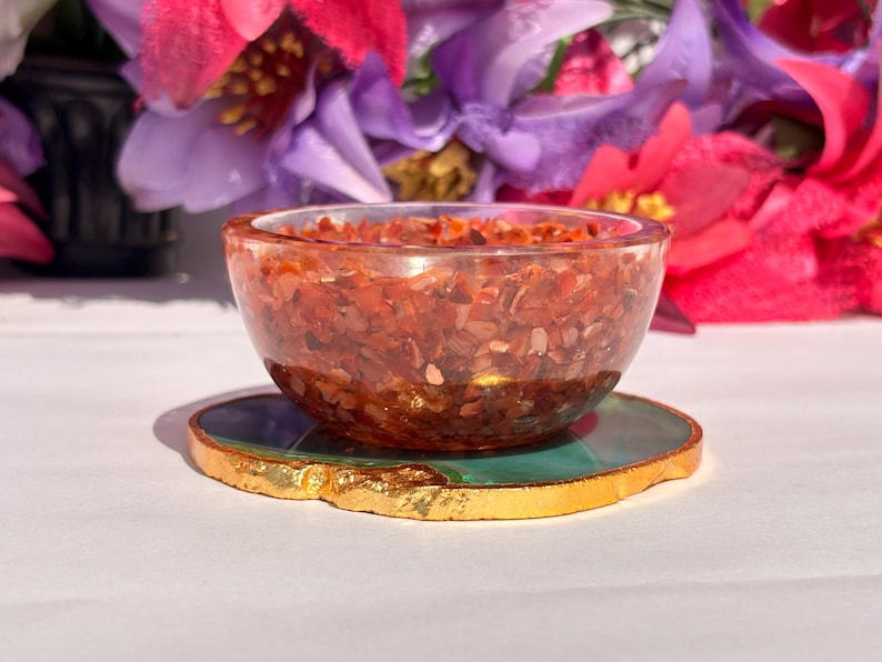 May include: A small, clear bowl filled with red, orange, and gold gemstone chips. The bowl is sitting on a green and gold agate coaster.