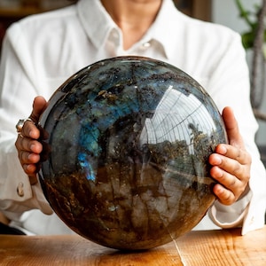 Könnte beinhalten: Eine große, polierte Labradoritkugel mit schillernden blauen und grünen Reflexen. Die Kugel wird mit beiden Händen gehalten und zeigt ihre glatte, reflektierende Oberfläche und natürlichen Muster. Die Kugel liegt auf einer Holzoberfläche.