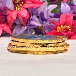 May include: Four round agate coasters with gold leaf accents. The coasters are stacked on top of each other. The coasters have a green, blue, and white marbled pattern.