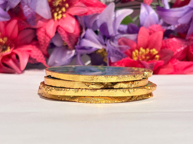 May include: Four round agate coasters with gold leaf accents. The coasters are stacked on top of each other. The coasters have a green, blue, and white marbled pattern.