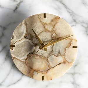 May include: A circular wall clock with a natural stone face. The clock face has beige, cream, and brown tones with gold-coloured hands and markers. The clock's hands are thin and elegant, and the markers are small rectangles.
