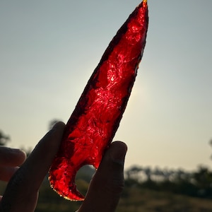 Puede incluir: Una punta de flecha de obsidiana roja con una punta afilada y una base curva. La punta de flecha es translúcida y tiene un ligero tono naranja.