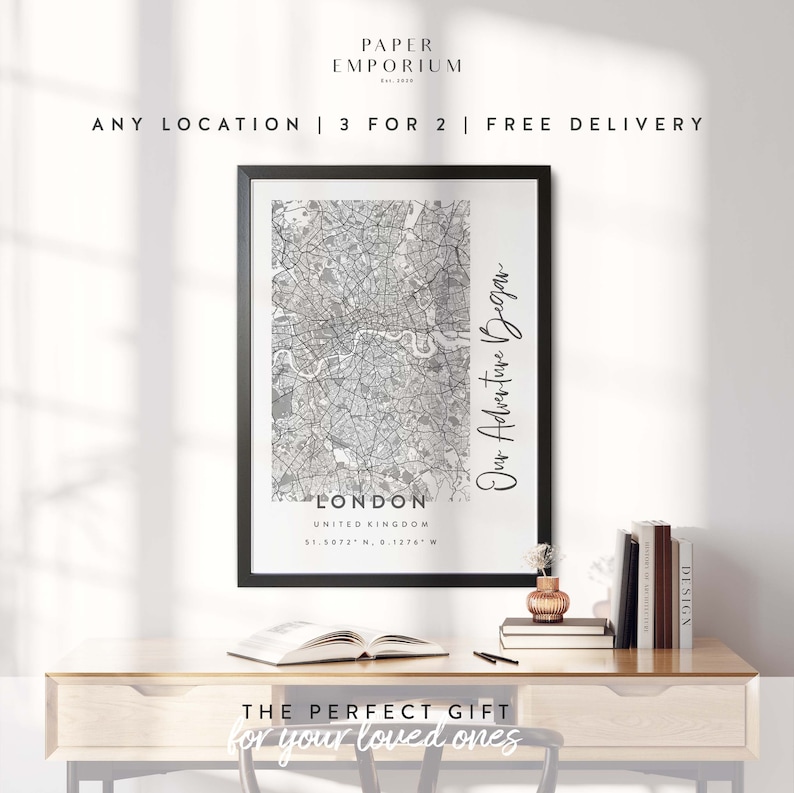 May include: A black and white map print of London, United Kingdom with the coordinates 51.5072&deg; N, 0.1276&deg; W. The text "Our Adventure Began" is written vertically on the right side of the map. The print is framed and displayed on a wooden desk with a vase of flowers and books.