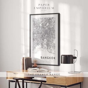 May include: Black and white map print of Bangkok, Thailand, in a black frame, hanging on a white wall above a wooden desk with a lamp and books. The print includes the coordinates 13.7563&deg; N 100.5018&deg; E and the text "THE FRAMED RANGE".