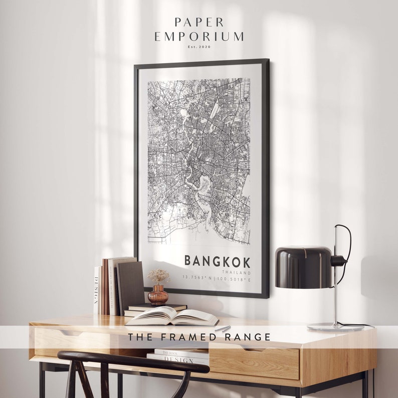 May include: Black and white map print of Bangkok, Thailand, in a black frame, hanging on a white wall above a wooden desk with a lamp and books. The print includes the coordinates 13.7563&deg; N 100.5018&deg; E and the text "THE FRAMED RANGE".