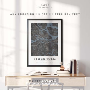 May include: A framed black and white map of Stockholm, Sweden with the city name and coordinates printed below the map. The map is on a wooden desk with a book, a vase with a small plant, and a stack of books.