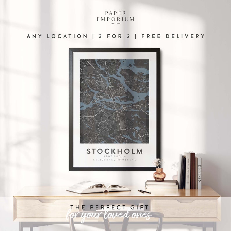 May include: A framed black and white map of Stockholm, Sweden with the city name and coordinates printed below the map. The map is on a wooden desk with a book, a vase with a small plant, and a stack of books.