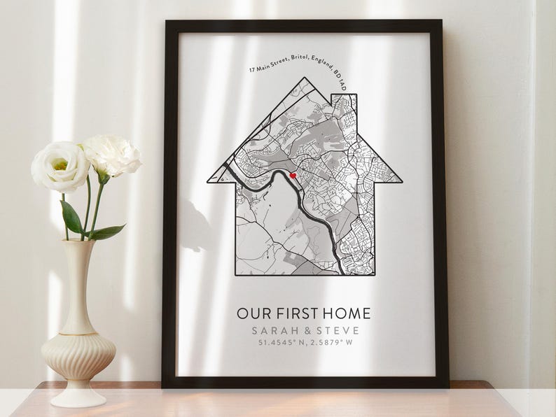 Op de afbeelding: Een zwart ingelijst afdruk met een witte achtergrond met een kaart van een wijk in de vorm van een huis. De tekst "Our First Home" is onder de kaart gedrukt, samen met de namen "Sarah & Steve" en de co&ouml;rdinaten "51.4545&deg; N, 2.5879&deg; W".