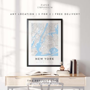 May include: A black framed print of a map of New York City with the text "New York, United States, 40.7128&deg;N, 74.0060&deg;W" below the map. The print is on a wooden desk with a book, a vase with flowers, and a stack of books.