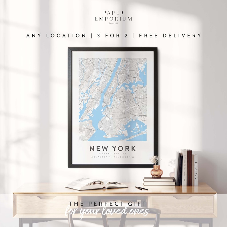 May include: A black framed print of a map of New York City with the text "New York, United States, 40.7128&deg;N, 74.0060&deg;W" below the map. The print is on a wooden desk with a book, a vase with flowers, and a stack of books.