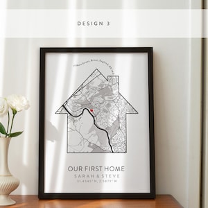 Op de afbeelding: Een zwart ingelijst afdruk met een witte achtergrond met een kaart van een wijk in de vorm van een huis. De tekst "OUR FIRST HOME" is onder de kaart gedrukt, samen met de namen "SARAH & STEVE" en de co&ouml;rdinaten "51.4545" N, 2.5879" W.