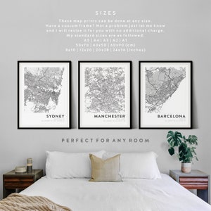 May include: Three black framed city maps of Sydney, Manchester, and Barcelona. The maps are printed in black and white and are hung on a white wall above a white bed with a beige throw blanket. The text "SIZES" is at the top of the image, followed by information about the sizes available. The text "PERFECT FOR ANY ROOM" is below the maps.