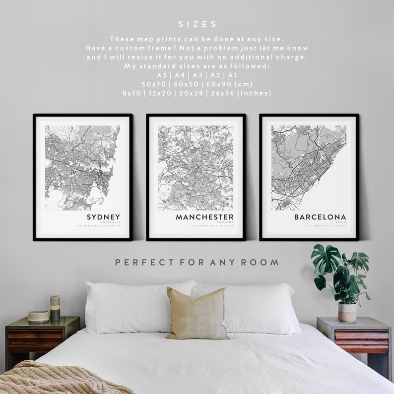 May include: Three black framed city maps of Sydney, Manchester, and Barcelona. The maps are printed in black and white and are hung on a white wall above a white bed with a beige throw blanket. The text "SIZES" is at the top of the image, followed by information about the sizes available. The text "PERFECT FOR ANY ROOM" is below the maps.
