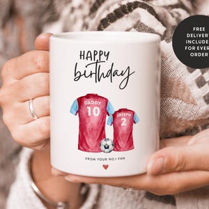 Op de afbeelding: Witte keramische mok met een rood en blauw aquarelontwerp van twee voetbalshirts met de woorden "Happy birthday", "Daddy 10", "Joseph 2" en "From your no.1 fan".