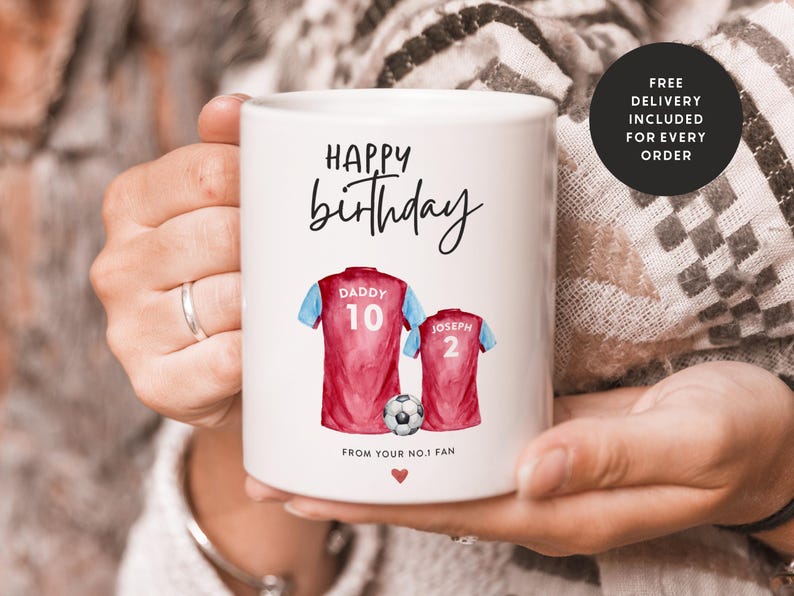 Op de afbeelding: Witte keramische mok met een rood en blauw aquarelontwerp van twee voetbalshirts met de woorden "Happy birthday", "Daddy 10", "Joseph 2" en "From your no.1 fan".