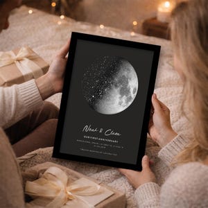May include: A framed black and white print featuring a detailed depiction of the moon and stars. The print includes the names "Noah & Clara", the text "Our First Anniversary", and other details. The overall theme is romantic and celestial.
