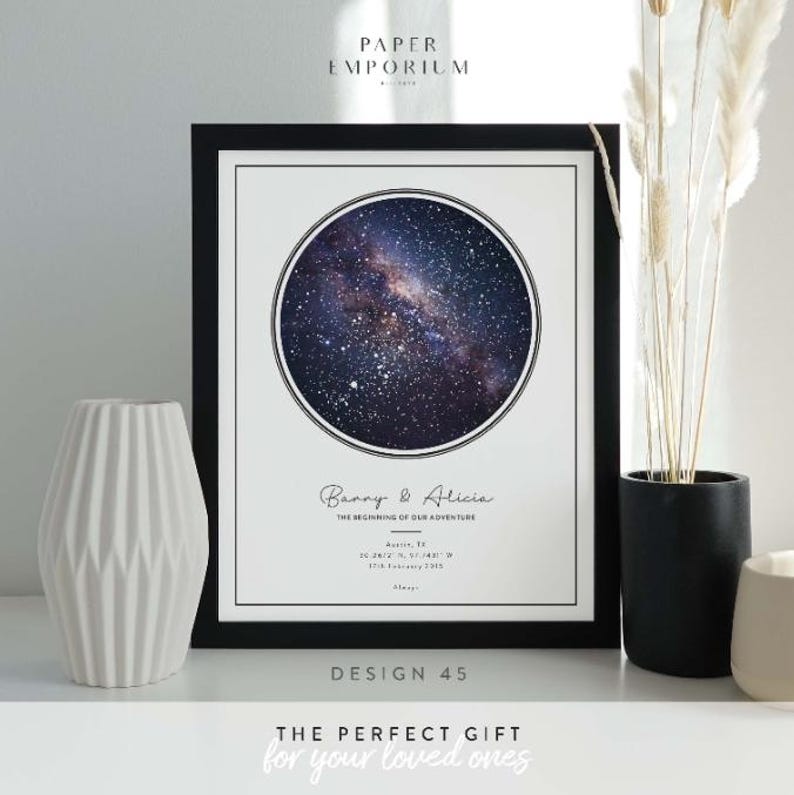May include: Framed print featuring a circular image of a starry galaxy. The print includes the names "Banny & Alicia" and the text "THE BEGINNING OF OUR ADVENTURE." The frame is black, and the print is displayed with a white vase and decorative plants.