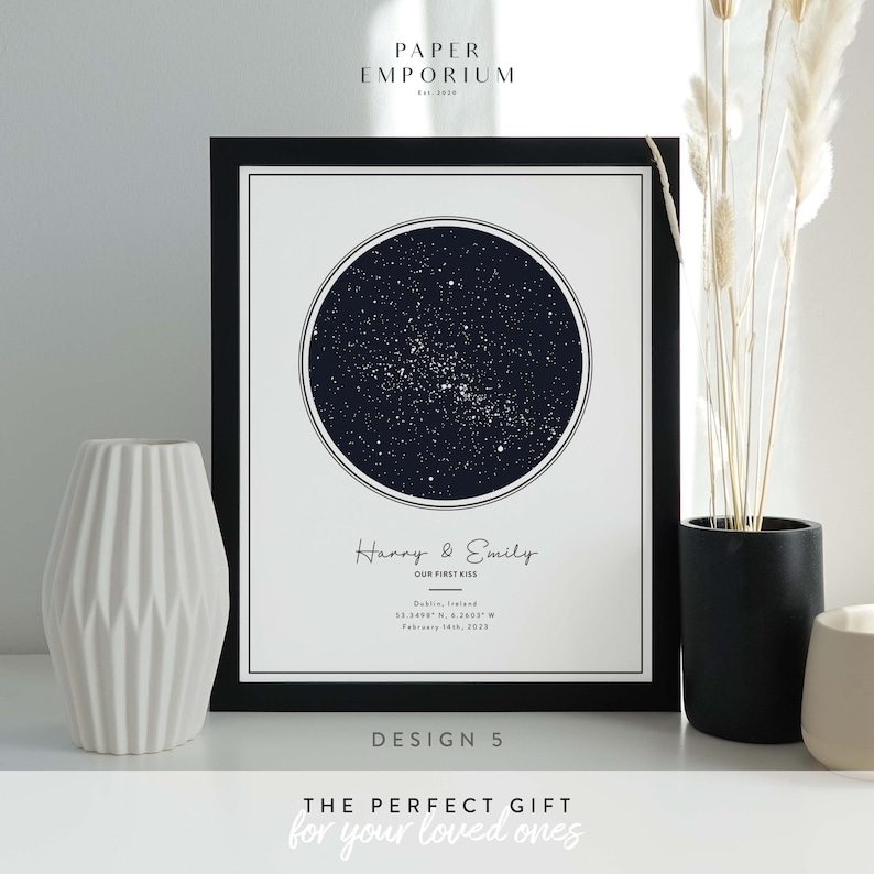 Op de afbeelding: Een zwart-witte sterrenkaartprint met de namen "Harry & Emily" en de tekst "Our First Kiss" samen met de locatie "Dublin, Ierland" en co&ouml;rdinaten "53.3498 N, 6.2603 W" en de datum "14 februari 2023". De print is ingelijst in een zwart frame en heeft de tekst "DESIGN 5" onder de kaart.
