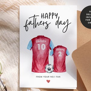 Op de afbeelding: Een witte kaart met de tekst "HAPPY, fathers day" in zwarte cursieve letters. Twee aquarel illustraties van voetbalshirts met de nummers 10 en 2, een met het label "DADDY" en de andere met "JOSEPH". Een voetbal ligt tussen de shirts. De kaart bevat ook de tekst "FROM YOUR NO.1 FAN" en een klein rood hart.
