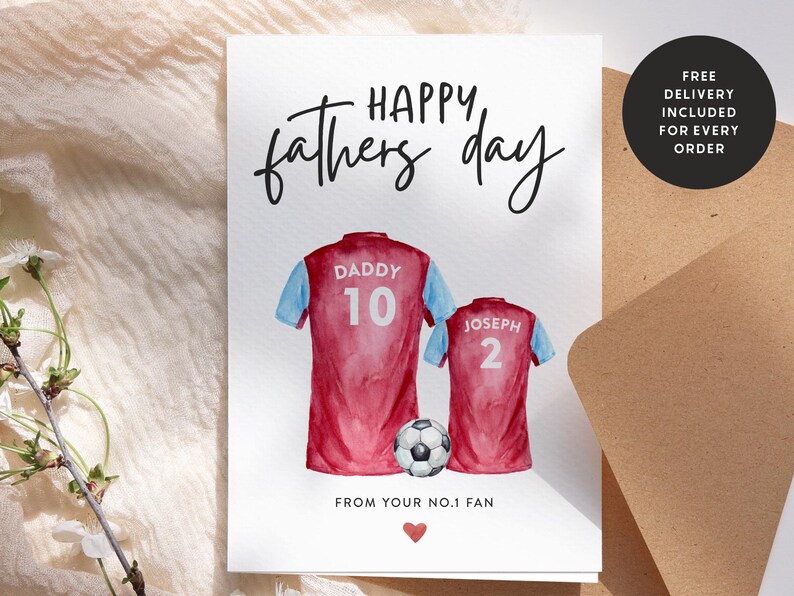 Op de afbeelding: Een witte kaart met de tekst "HAPPY, fathers day" in zwarte cursieve letters. Twee aquarel illustraties van voetbalshirts met de nummers 10 en 2, een met het label "DADDY" en de andere met "JOSEPH". Een voetbal ligt tussen de shirts. De kaart bevat ook de tekst "FROM YOUR NO.1 FAN" en een klein rood hart.