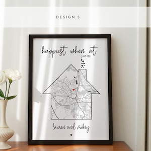 Op de afbeelding: Een zwart ingelijst afdruk met de tekst "Happiest when at home" en een kaart van een stad met een rood hart in het midden. De tekst "Lauren and Mikey" staat onderaan de afdruk.