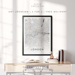 May include: A black and white map print of London, England, in a black frame. The map shows the city's streets and landmarks. The print is on a wooden desk with a book and a vase of flowers. The text "THE PERFECT GIFT for your loved ones" is below the print.
