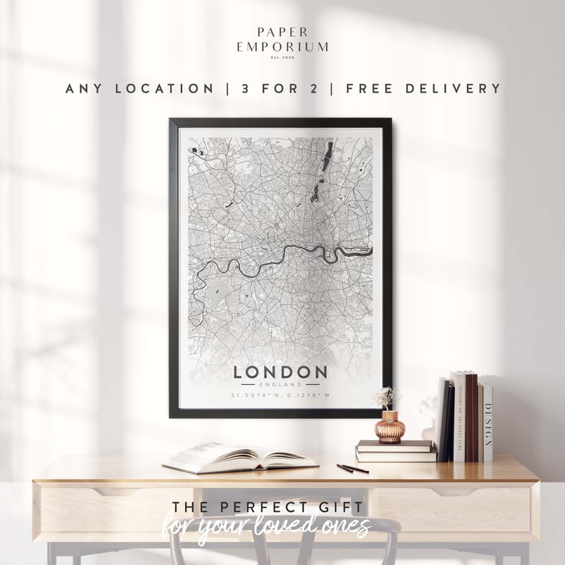 May include: A black and white map print of London, England, in a black frame. The map shows the city's streets and landmarks. The print is on a wooden desk with a book and a vase of flowers. The text "THE PERFECT GIFT for your loved ones" is below the print.