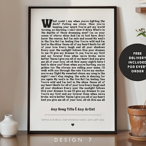 May include: A framed print with song lyrics in black font on a white background. The print includes the text "Any Song Title & Any Artist" and a wedding date. A small white vase and a potted plant are visible.
