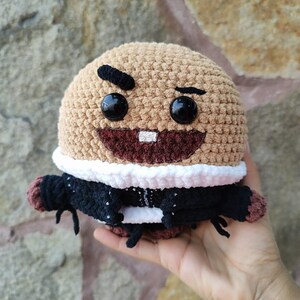Puede incluir: Muñeca de ganchillo hecha a mano con cabeza color canela, cejas negras y ojos negros grandes. La muñeca tiene una boca ancha y sonriente con un solo diente. Lleva una chaqueta negra con ribete blanco y brazos negros pequeños.