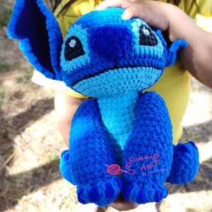 Könnte beinhalten: Ein blaues, gehäkeltes Stitch-Plüschtier mit einem großen Lächeln und großen Ohren. Das Spielzeug wird von einem Kind gehalten.